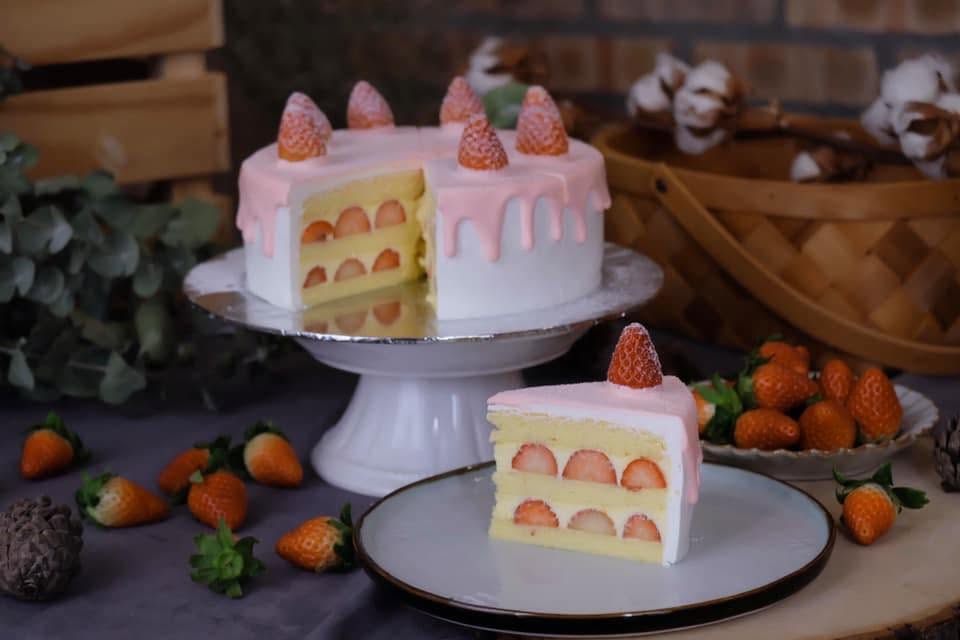 A whole strawberry cream cake with a soft pink drip and fresh strawberries, beside a cut slice showing layered sponge — styled with eucalyptus on a rustic table
