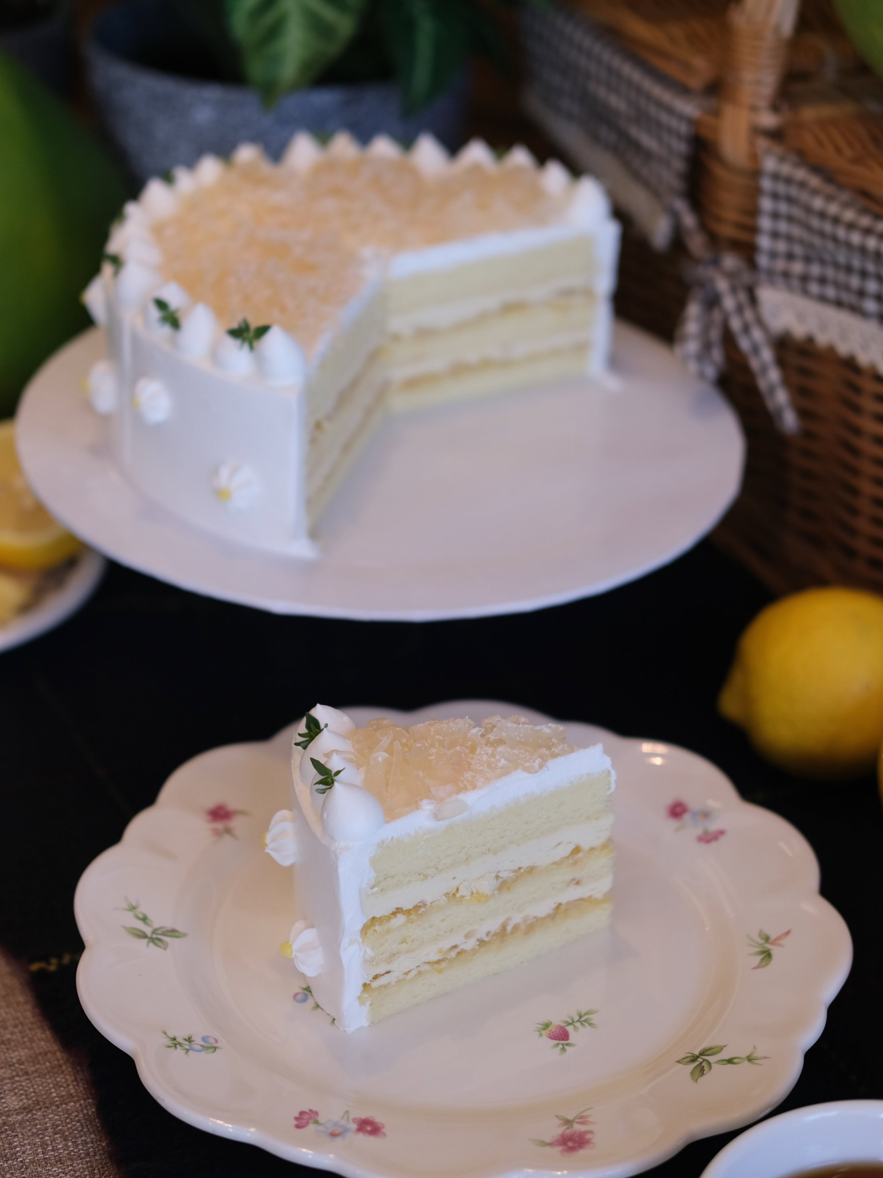 A homey layered lemon cream cake — pale sponge with whipped cream layers and a candied-lemon crown, a slice on a floral plate beside fresh lemons