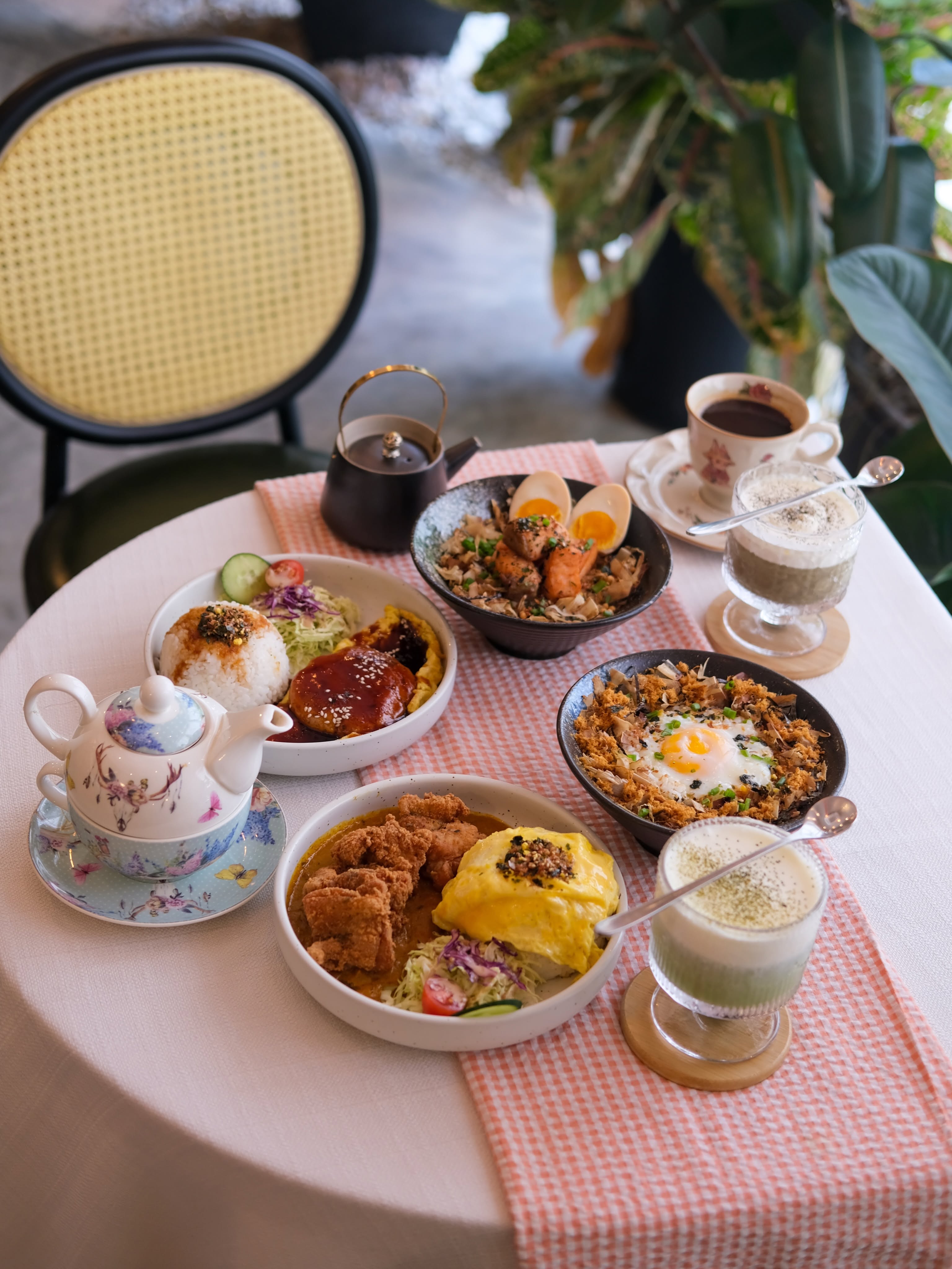 An all-day brunch spread at Dreamz Bakery — rice bowls with runny egg, fried chicken, sides, a pot of tea, matcha and coffee lattes on a pink-striped table by a cane chair and plants