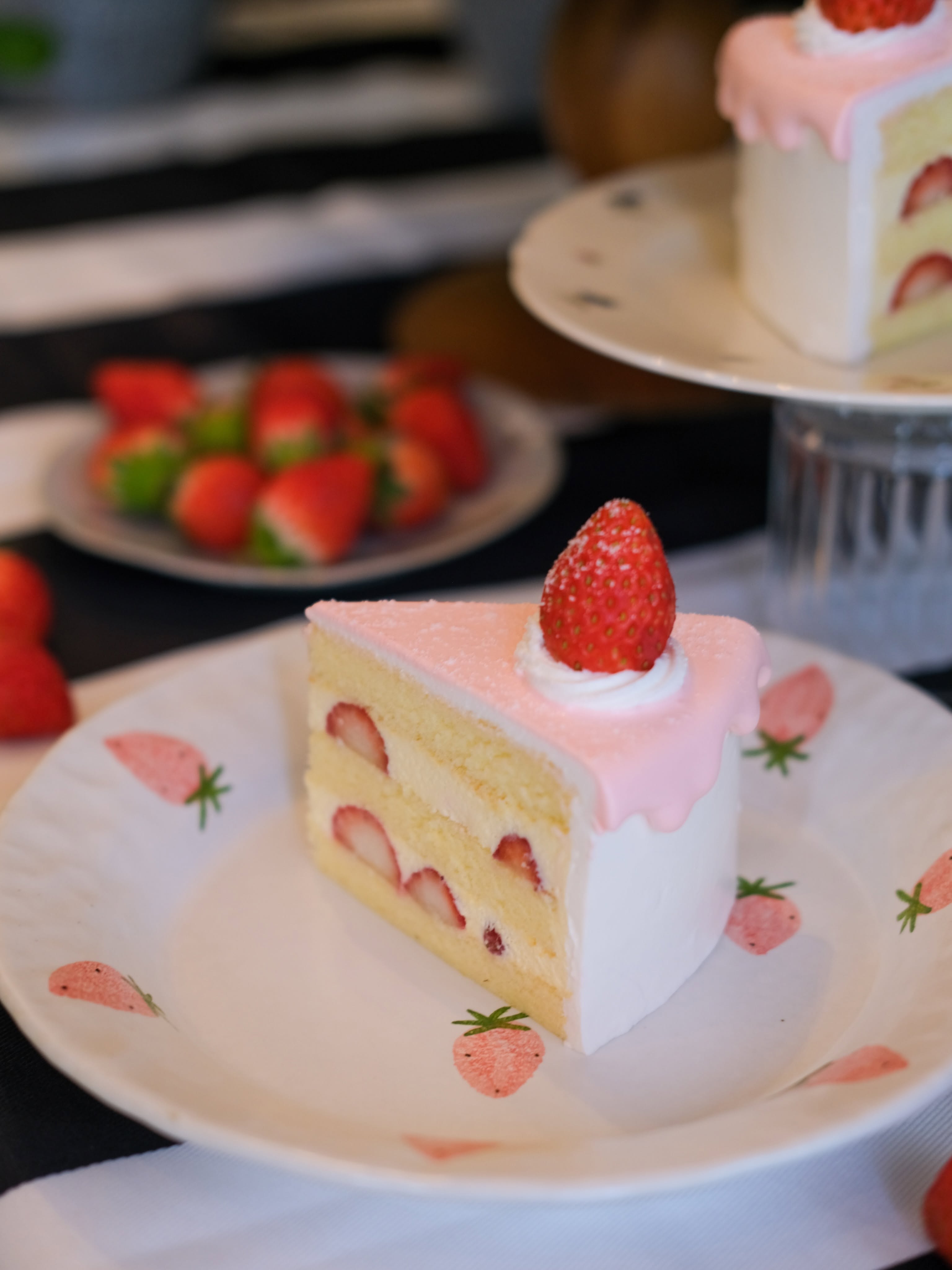 A slice of strawberry shortcake — layers of sponge and fresh cream studded with strawberries, topped with a soft pink drip and a whole dusted strawberry, on a strawberry-print plate